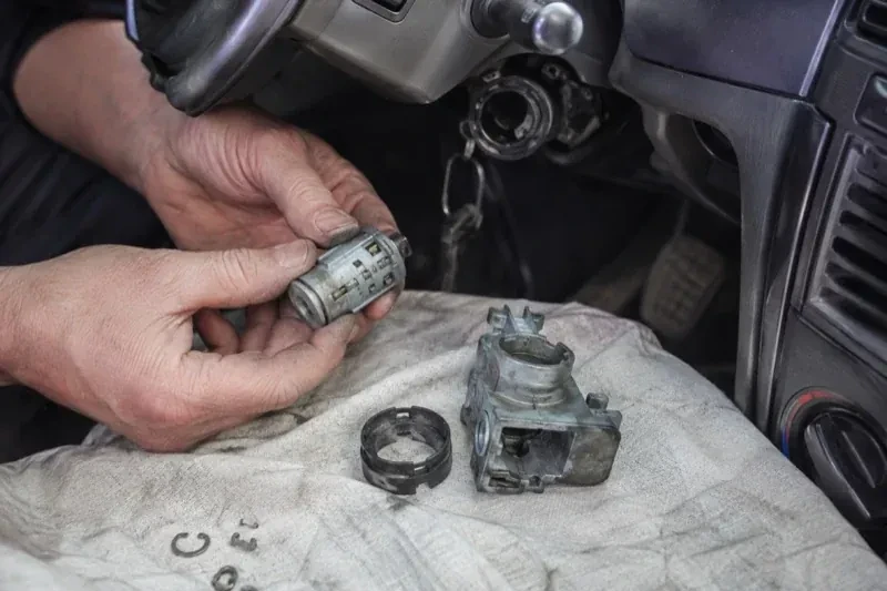 Hands holding a removed ignition cylinder beside a car steering column, with the housing and small parts on a cloth. — Ignition Lock Repair, Norfolk County